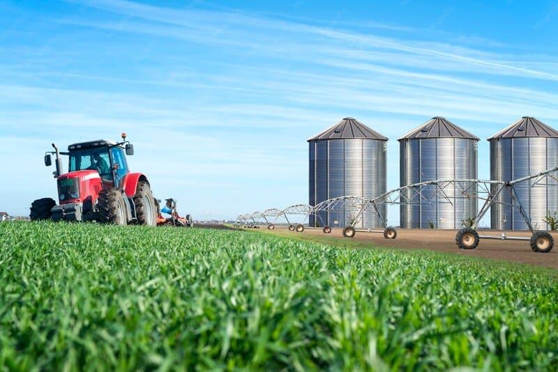 Red tractor working a field beside grain silos and irrigation equipment